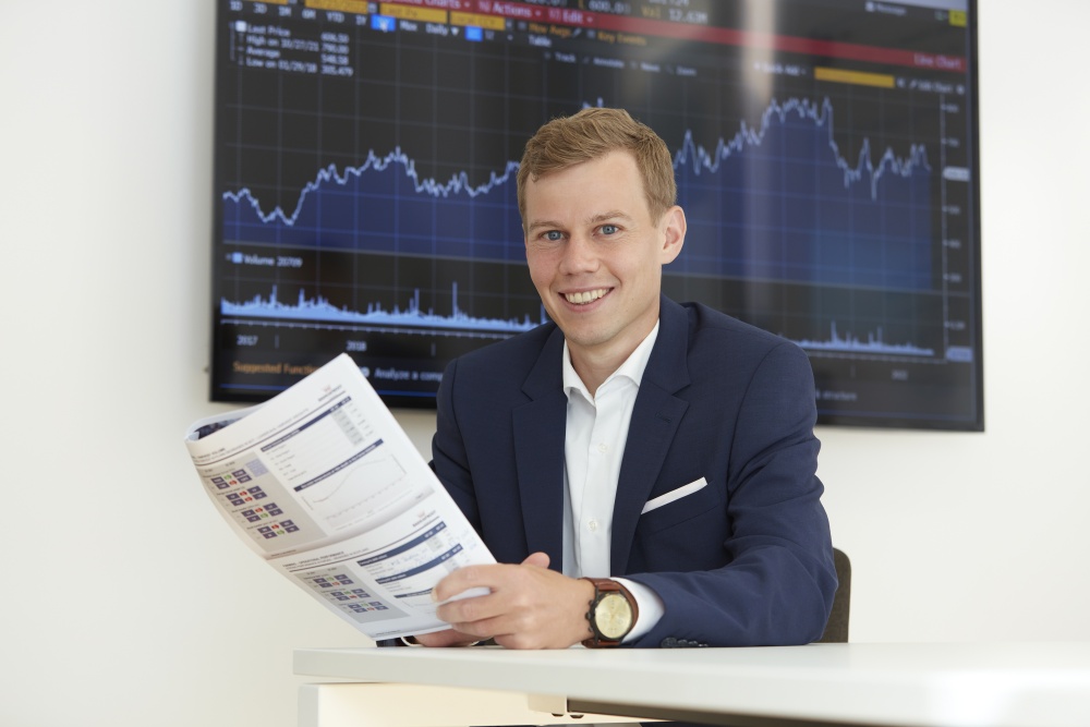 Philipp Hämmerli im Businessanzug hält einen Finanzbericht in der Hand und sitzt vor einem Bildschirm mit Aktienkurs- und Marktgrafiken.
